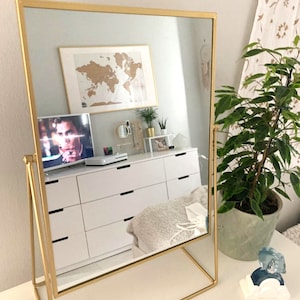 Peut inclure: Un miroir sur pied avec un cadre doré, un meuble blanc et une télévision reflétés dans le verre. Le miroir est sur un pied en métal doré de forme triangulaire.