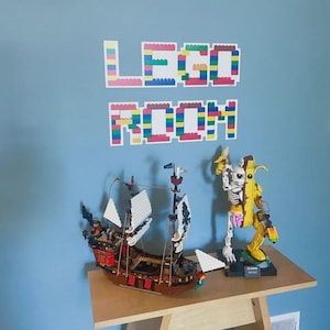 May include: A brown Lego pirate ship with white sails and red accents sits on a wooden table. Beside it is a Lego figure of a skeleton inside a yellow banana suit. The words "LEGO ROOM" are spelled out on the blue wall above.
