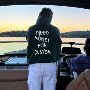 Op de afbeelding: Een persoon met een donkergroene hoodie met de witte tekst "NEED MONEY FOR CUSTOM" staat op een boot. De persoon staat met de rug naar de camera. Een andere persoon bestuurt de boot. De achtergrond toont een meer en bomen.