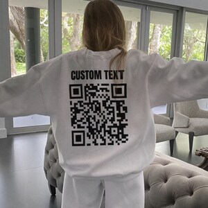 May include: White sweatshirt with a black QR code and the text "CUSTOM TEXT" printed on the back.