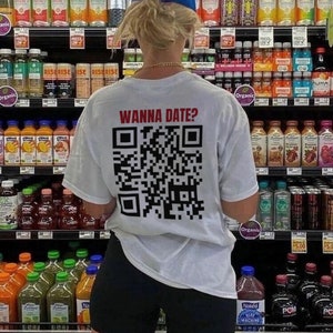 Puede incluir: Una camiseta blanca con un código QR en blanco y negro en la espalda. El texto "Wanna Date?" está impreso encima del código QR. La camiseta la lleva puesta una persona en una tienda de comestibles.