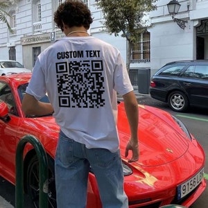 Pode incluir: Uma camiseta branca com um código QR preto e o texto "CUSTOM TEXT" impresso nas costas. A pessoa que está usando a camiseta está ao lado de um carro esportivo vermelho.