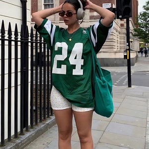 Può includere: Una maglia da football verde e bianca con il numero 24 in bianco. La maglia è abbinata a pantaloncini bianchi con un motivo floreale. Una borsa tote verde è portata a tracolla. La persona indossa occhiali da sole e cuffie.