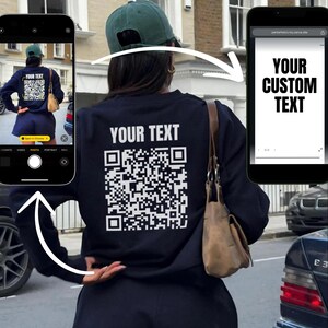 May include: A black sweatshirt with a white QR code and the text "YOUR TEXT" printed on the back. The sweatshirt is being worn by a person with a black hat and a brown bag.
