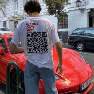 Könnte beinhalten: Ein weißes T-Shirt mit dem Text "Wanna Date Me?" und einem schwarz-weißen QR-Code auf der Rückseite. Die Person, die das Shirt trägt, steht neben einem roten Sportwagen.