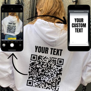 Op de afbeelding: Witte hoodie met een zwarte QR-code en de tekst "YOUR TEXT" gedrukt op de rug. De hoodie wordt gedragen door iemand die blauwe jeans draagt.