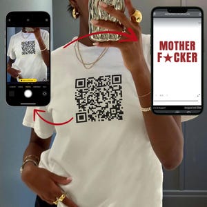 Può includere: T-shirt bianco con un grande codice QR nero e la scritta "MOTHER F*CKER" in rosso. L'immagine mostra una persona che tiene in mano un telefono che mostra lo stesso testo. La maglietta a maniche corte sembra essere di cotone.