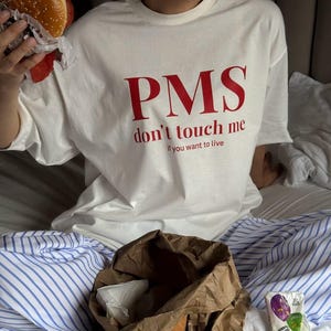 Könnte beinhalten: Ein weißes kurzärmeliges T-Shirt mit dem roten Aufdruck PMS don't touch me if you want to live auf der Brust. Das T-Shirt wird mit einer blau-weiß gestreiften Schlafanzughose kombiniert, was einen lässigen Look für entspannte Stunden zu Hause ergibt.