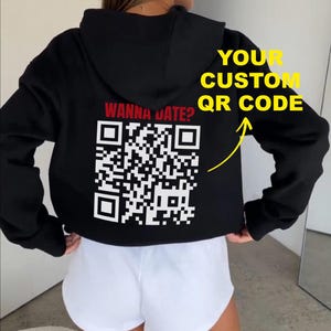 Op de afbeelding: Een zwarte hoodie met de rode tekst WANNA DATE? boven een grote witte QR-code op de achterkant. Het ontwerp bevat een gele pijl die naar de QR-code wijst met de gele tekst YOUR CUSTOM QR CODE ernaast.