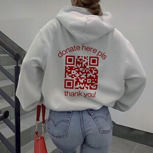 Puede incluir: Sudadera blanca con un código QR rojo y el texto "donate here pls thank you!" impreso en la espalda.