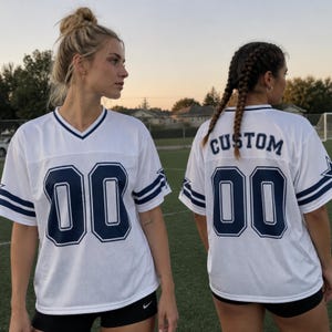 May include: A white short-sleeved football jersey with a V-neck collar. The front features the number 00 in navy blue with a white outline. The back displays the word CUSTOM above the number 00 in navy blue. Navy blue stripes accent the sleeves.