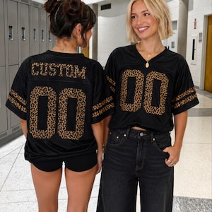 May include: A black jersey with short sleeves featuring a leopard print pattern on the numbers and stripes. The back displays the word CUSTOM in leopard print above the number 00, while the front features the number 00 in the same print.