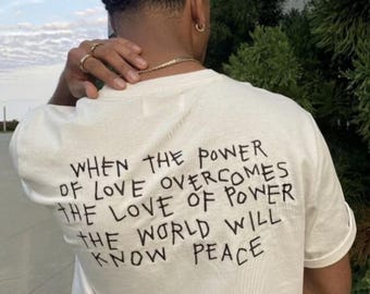 以下が含まれることがあります： 「WHEN THE POWER OF LOVE OVERCOMES THE LOVE OF POWER THE WORLD WILL KNOW PEACE」と黒い文字で書かれたクリーム色のTシャツ。 金色のジュエリーと金のチェーンを身に着けている。