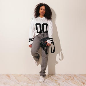 Puede incluir: Una persona lleva una camiseta blanca de manga larga con el n&uacute;mero "00" en negro en la parte delantera. La camiseta tiene rayas negras en las mangas. La persona lleva vaqueros grises, zapatillas blancas y un bolso bandolera negro.