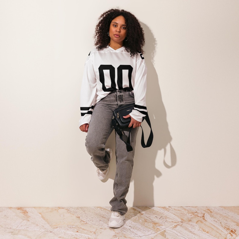 Puede incluir: Una persona lleva una camiseta blanca de manga larga con el n&uacute;mero "00" en negro en la parte delantera. La camiseta tiene rayas negras en las mangas. La persona lleva vaqueros grises, zapatillas blancas y un bolso bandolera negro.