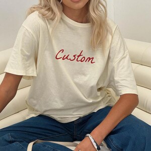 May include: Cream-colored t-shirt with the word "Custom" in red script. The shirt has short sleeves and a relaxed fit. The person is wearing blue jeans and a silver bracelet.