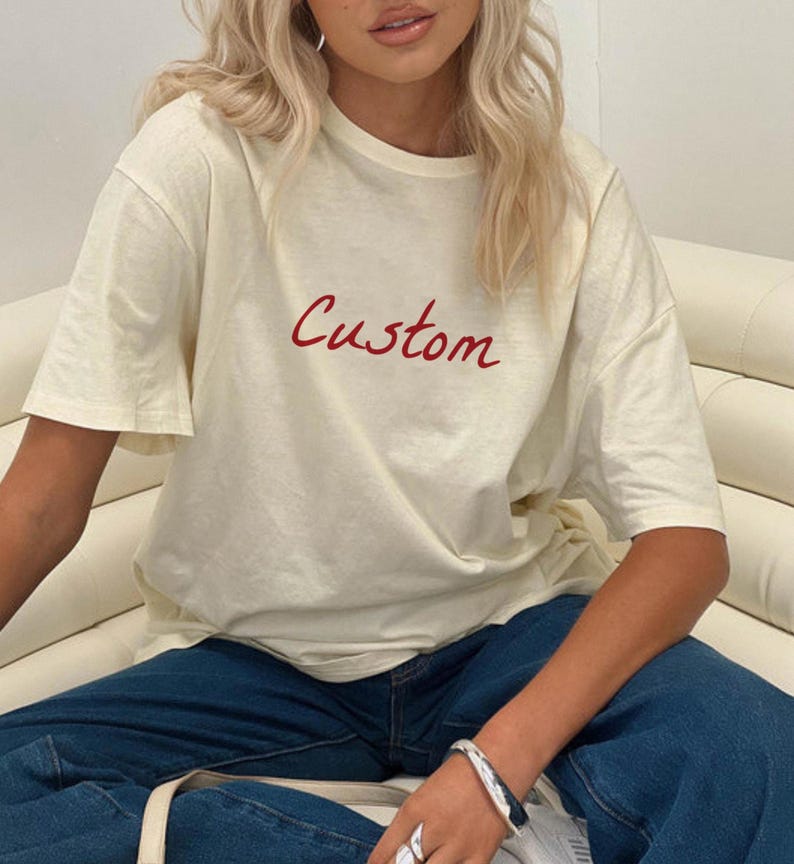 May include: Cream-colored t-shirt with the word "Custom" in red script. The shirt has short sleeves and a relaxed fit. The person is wearing blue jeans and a silver bracelet.