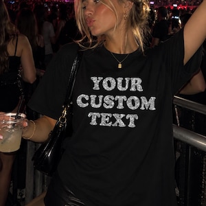 May include: A black short-sleeved t-shirt featuring the words YOUR CUSTOM TEXT printed in a silver glitter texture across the chest. The garment has a crew neckline and a relaxed fit, providing a simple base for personalized graphic designs.