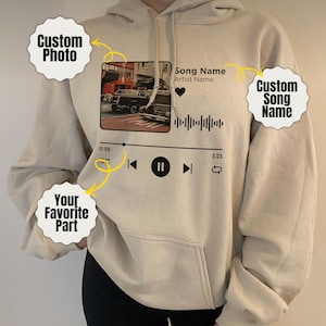 May include: A cream-coloured hoodie featuring a custom photo and song graphic. The graphic includes a photo, song name, artist name, and a music player interface with play, pause, and skip buttons. The hoodie has a front pocket and a drawstring hood.