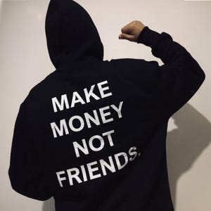 Könnte beinhalten: Schwarzer Kapuzenpullover mit dem weißen Text "MAKE MONEY NOT FRIENDS" aufgedruckt. Auf dem Bild ist die Rückseite des Kapuzenpullovers zu sehen, mit hochgezogener Kapuze. Die Person, die den Kapuzenpullover trägt, beugt ihren Arm.