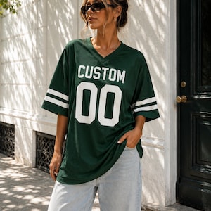 Puede incluir: Una camiseta deportiva de color verde oscuro con cuello en V y mangas cortas que presentan dos rayas horizontales blancas. El frente de la prenda muestra la palabra CUSTOM en letras blancas sobre los números 00 en un estilo de fuente bloque.