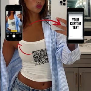 Op de afbeelding: Een wit tanktop met een zwart-wit QR-code ontwerp. Het tanktop wordt vastgehouden door een persoon die een blauw-wit geruit overhemd draagt. De tekst "YOUR CUSTOM TEXT" is zichtbaar op een telefoonscherm.