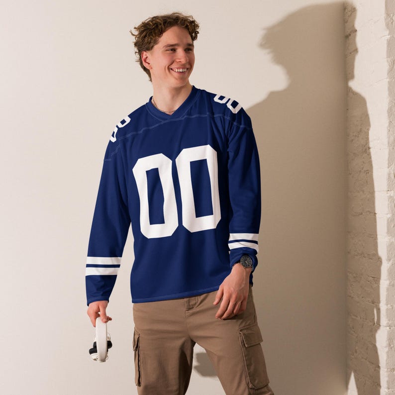 Puede incluir: Una camiseta de f&uacute;tbol de manga larga azul marino con el n&uacute;mero "00" en blanco en la parte delantera. La camiseta tiene rayas blancas en las mangas y los hombros. La persona lleva pantalones cargo color caqui y un reloj.