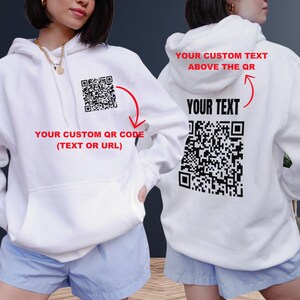 May include: White hoodie with a black and white QR code on the front and back. The text "YOUR CUSTOM QR CODE (TEXT OR URL)" is printed below the QR code on the front. The text "YOUR CUSTOM TEXT ABOVE THE QR" is printed above the QR code on the back. The text "YOUR TEXT" is printed below the QR code on the back.