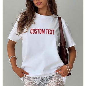 May include: A white t-shirt with the words "CUSTOM TEXT" printed in red. The shirt has short sleeves and a crew neck. The model is wearing a silver bracelet and a brown purse.