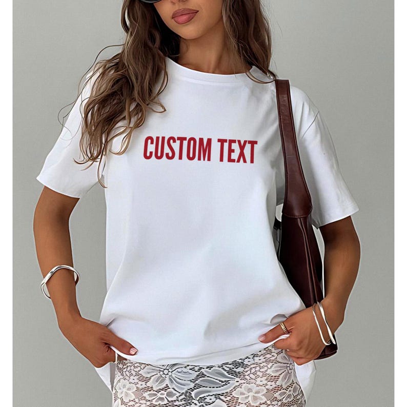 May include: A white t-shirt with the words "CUSTOM TEXT" printed in red. The shirt has short sleeves and a crew neck. The model is wearing a silver bracelet and a brown purse.