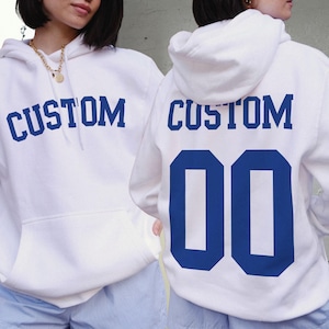Pu&ograve; includere: Due felpe con cappuccio bianche con testo e numeri blu. La felpa con cappuccio a sinistra ha "CUSTOM" stampato sul davanti. La felpa con cappuccio a destra ha "CUSTOM" stampato sul retro con il numero "00" sotto.