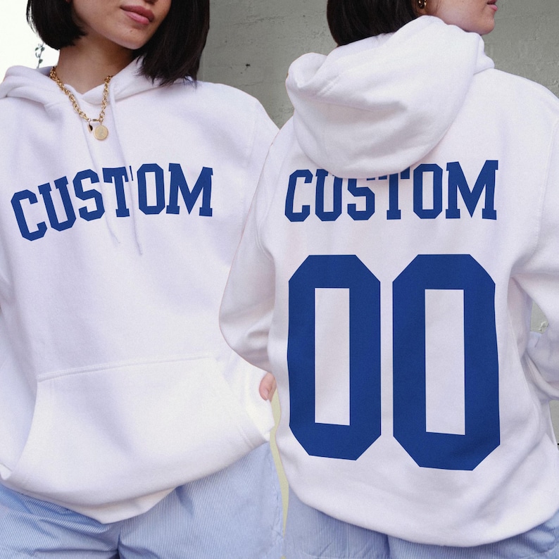 Pu&ograve; includere: Due felpe con cappuccio bianche con testo e numeri blu. La felpa con cappuccio a sinistra ha "CUSTOM" stampato sul davanti. La felpa con cappuccio a destra ha "CUSTOM" stampato sul retro con il numero "00" sotto.