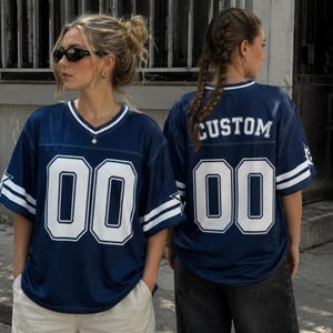 May include: A navy blue short-sleeved jersey with a V-neck collar, featuring white stripes on the sleeves and the number 00 in white on the front. The back displays the number 00 in white above the text CUSTOM in white block lettering.