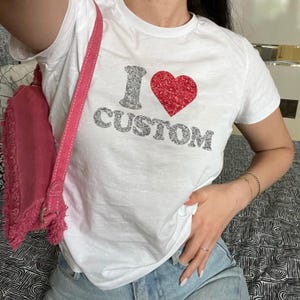 May include: A white short-sleeved t-shirt featuring the text I, a red glitter heart, and the word CUSTOM in silver glitter lettering. A pink suede shoulder bag with a fringed edge is positioned on the left side of the garment.