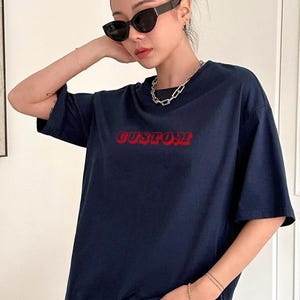 May include: Navy blue t-shirt with the word "CUSTOM" in red, bold, retro-style lettering. The shirt has short sleeves and a crew neck. The model is wearing black sunglasses and a silver chain necklace.