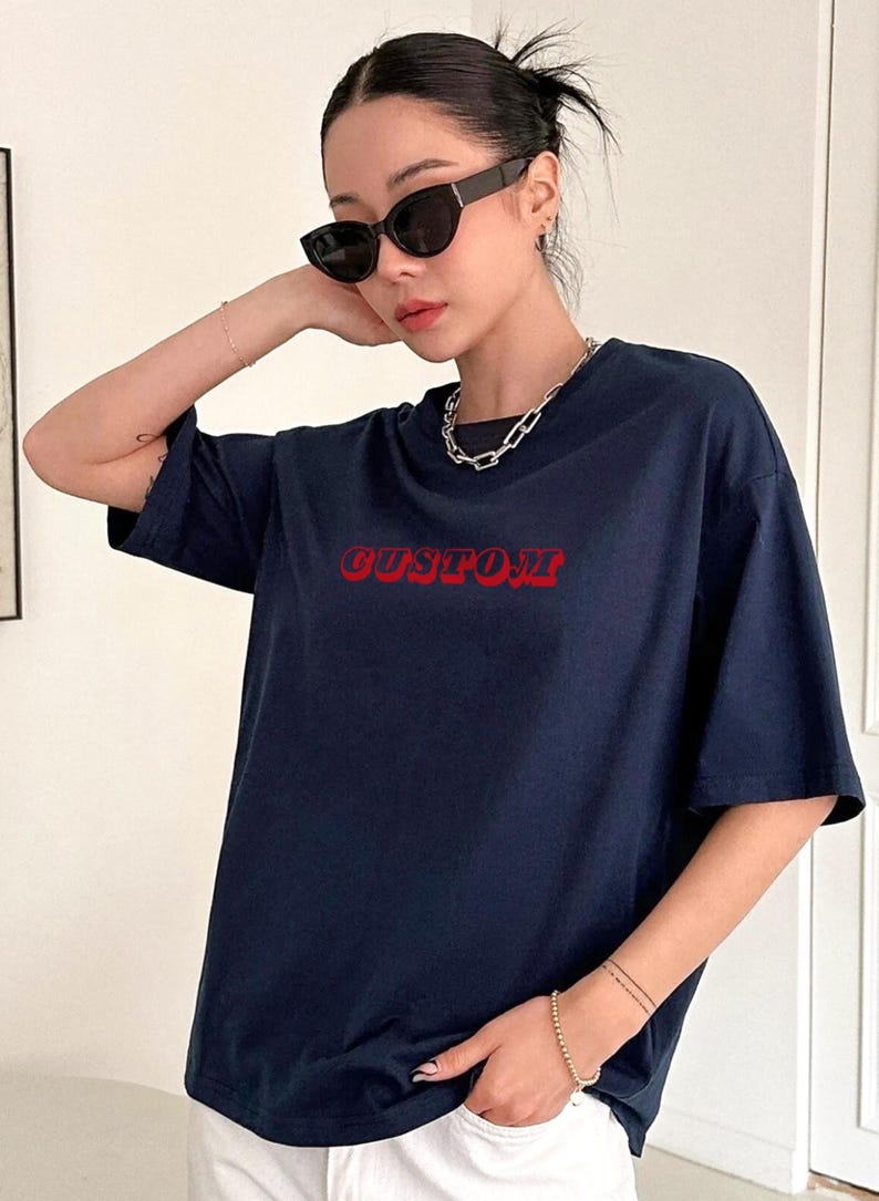 May include: Navy blue t-shirt with the word "CUSTOM" in red, bold, retro-style lettering. The shirt has short sleeves and a crew neck. The model is wearing black sunglasses and a silver chain necklace.
