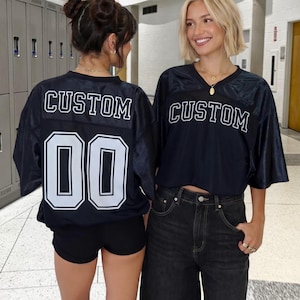 Puede incluir: Dos personas con camisetas de fútbol azul marino con la palabra "CUSTOM" en la espalda y el número "00". Las camisetas tienen letras y números blancos. Una persona lleva pantalones cortos negros, la otra vaqueros negros.