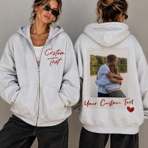 Peut inclure: Un sweat à capuche zippé gris clair avec le texte rouge Custom Text sur le devant. Le dos présente une impression photo rectangulaire de deux personnes s'enlaçant, avec le texte rouge Your Custom Text et un petit cœur rouge en dessous.