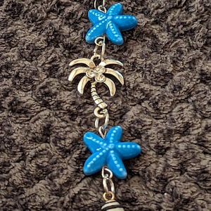 May include: A silver charm bracelet with blue star charms and palm tree charms. The palm tree charms have rhinestones. The bracelet ends with a black and white striped shell charm.