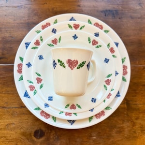 May include: A set of cream-colored ceramic dinnerware consisting of two stacked round plates and a matching mug. Each piece features a repeating pattern of red hearts, green leaves, and small blue diamond shapes arranged in a circular design.