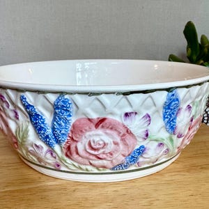 May include: White ceramic bowl with embossed floral design. The bowl features a pink rose, blue and purple flowers, and a woven basket pattern. The rim and base have green trim. A decorative piece for the home.