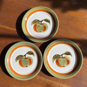 May include: Three ceramic plates with a white center, orange ring, and green rim. Each plate features a hand-painted apple design in green and orange. The plates are arranged on a wooden surface, showcasing a rustic aesthetic.