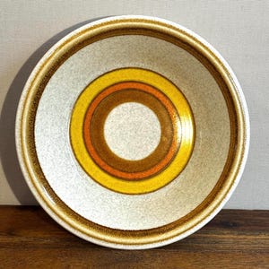 May include: A round ceramic bowl with a speckled white interior and a concentric circle design. The circles are yellow, orange, and brown, with a brown rim. The bowl is set on a wooden surface.