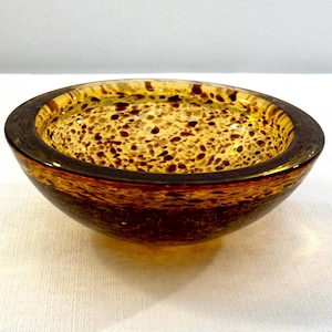 May include: A small, round glass bowl with a speckled interior. The bowl has a dark brown rim and a gradient of amber and brown colors. The interior is filled with small, dark brown spots, creating a unique pattern. The bowl is likely used for decorative purposes.