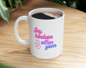 May include: White ceramic coffee mug with a pink and blue design that says "Say kocken offen yom" with a peace sign.