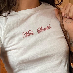 May include: White t-shirt with the embroidered text "Mrs. Smith" in a cursive font. The shirt is paired with blue jeans. A brown and tan handbag is held in the right hand.