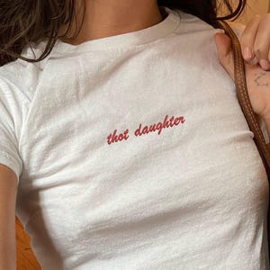 Puede incluir: Camiseta blanca con el texto "thot daughter" en escritura roja. La camiseta se combina con vaqueros azules. Se ve un bolso marrón con un estampado de monograma.