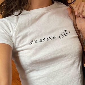 Pode incluir: Camiseta branca com o texto "it's no use, Jo!" estampado na frente. A camiseta está sendo usada por uma pessoa e está enfiada em uma calça jeans azul.