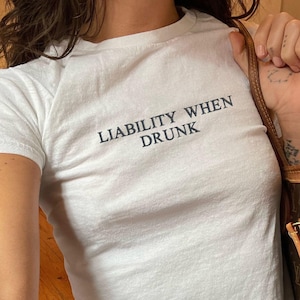 Könnte beinhalten: Weißes T-Shirt mit dem Text "LIABILITY WHEN DRUNK" in Dunkelblau. Das Shirt wird mit einer blauen Jeans kombiniert. Eine braune und beige Handtasche wird im Hintergrund gehalten.
