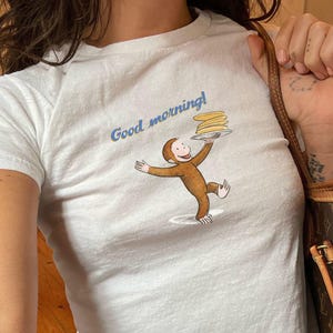 Op de afbeelding: Wit T-shirt met een afbeelding van een aap die een stapel pannenkoeken vasthoudt en de tekst "Good morning!" erop gedrukt.